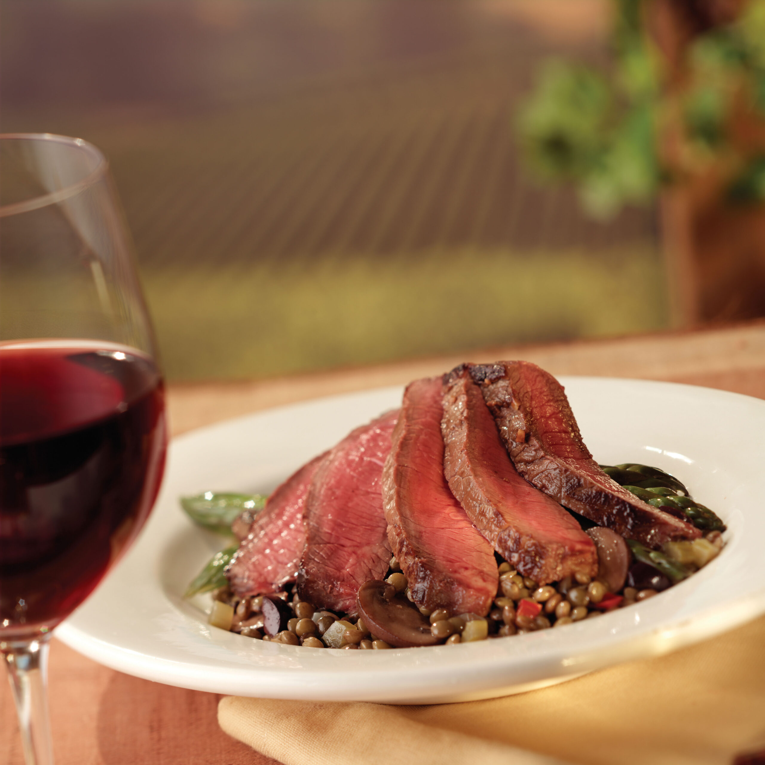 Tender slices of roasted beef served on a bed of lentils and sautéed vegetables, accompanied by a glass of red wine, set against a blurred vineyard background.