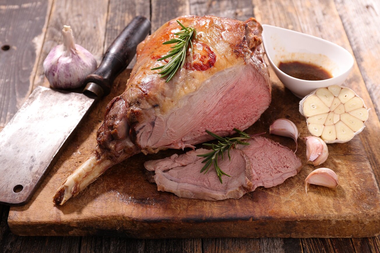 Roasted leg of lamb resting on a wooden cutting board, garnished with fresh rosemary and accompanied by garlic cloves and a small bowl of sauce, showcasing a perfectly cooked pink interior.