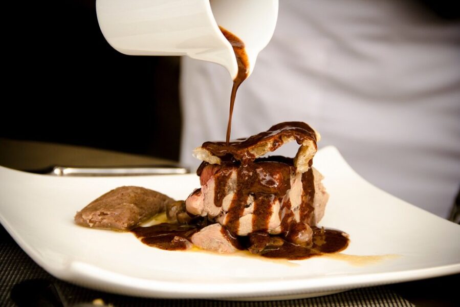 A chef pouring rich brown sauce over a beautifully plated dish of meat, showcasing gourmet presentation on a white plate.
