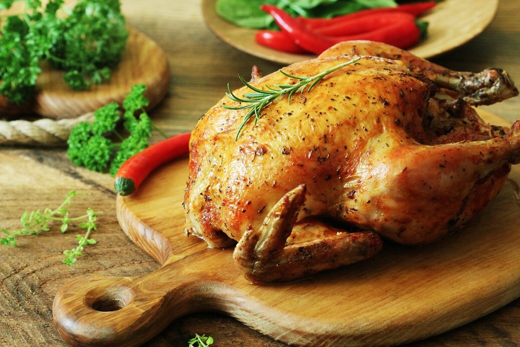 Roasted whole chicken garnished with fresh rosemary, served on a wooden cutting board, accompanied by red chili peppers and parsley, with a background of leafy greens. Perfect for a delicious dinner or special occasion.